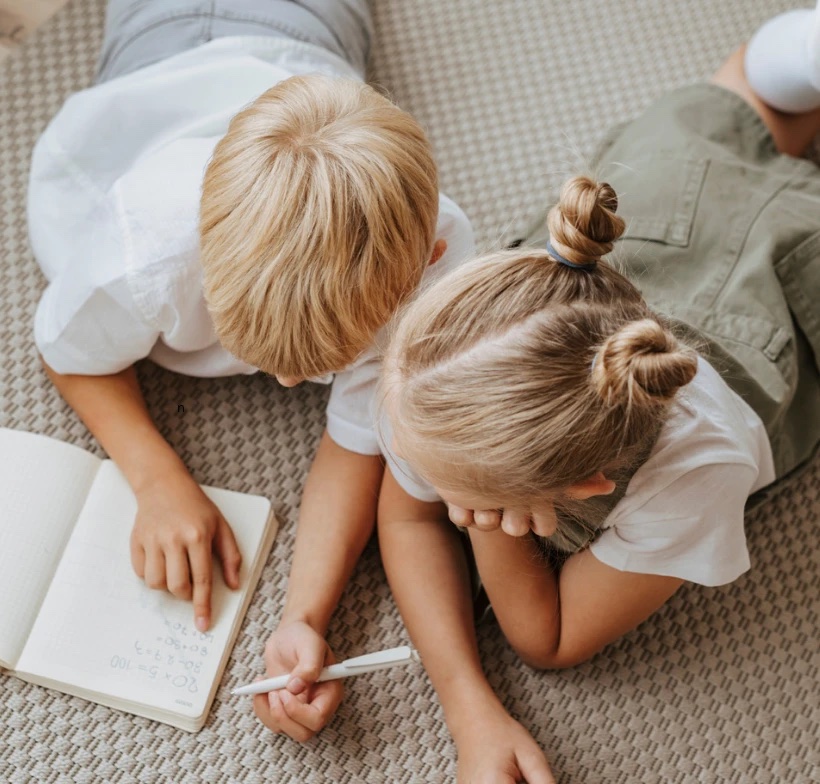 Two young children lying on the floor, writing in a notebook together